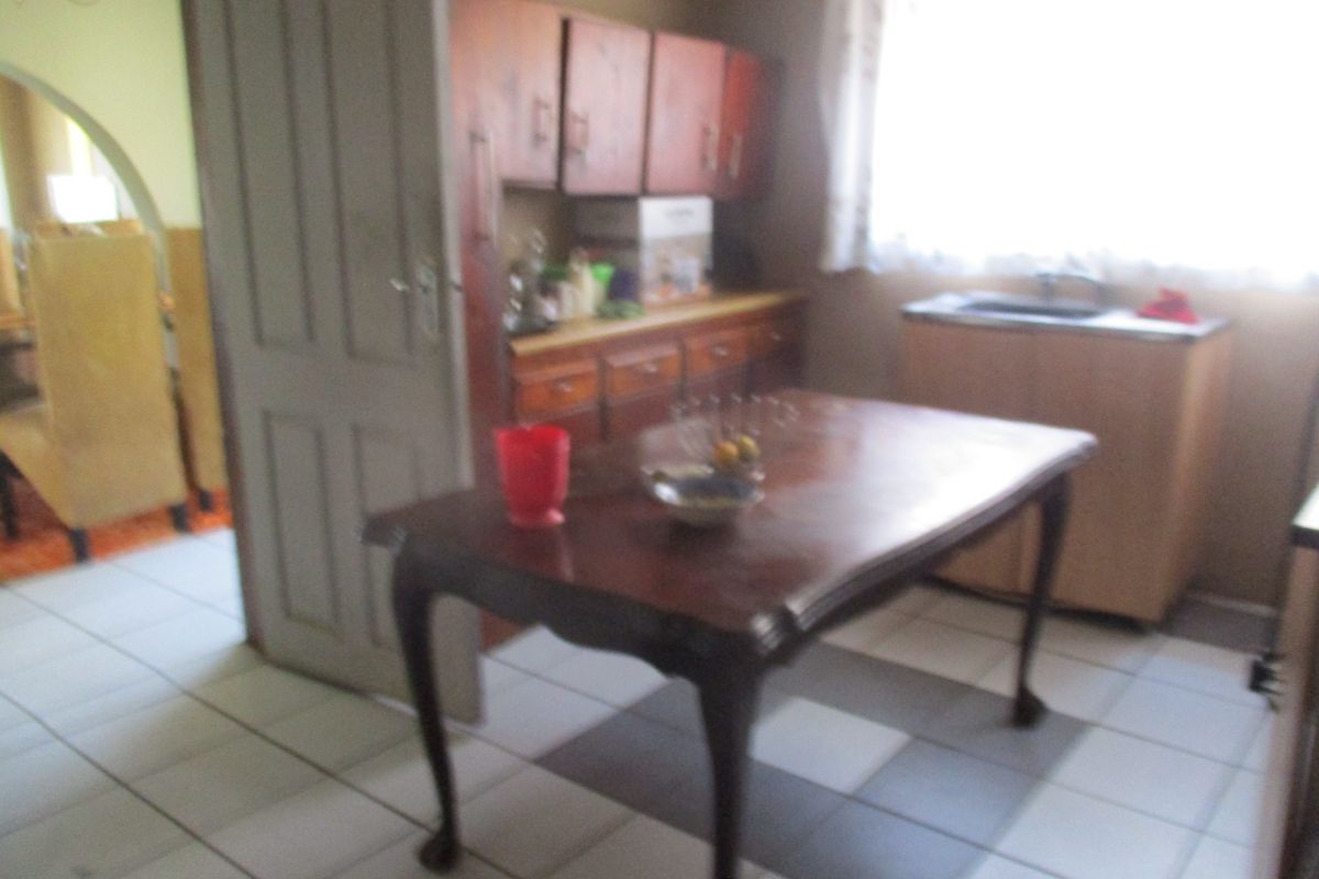A dated kitchen area featuring wooden cabinetry, a built-in sink, and tiled flooring, with an adjacent dining space visible through the doorway.