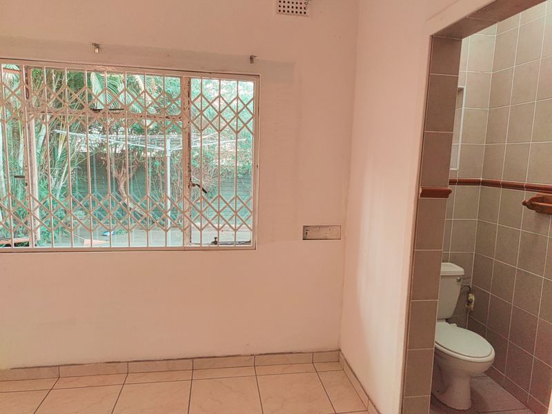 A view of an interior space featuring tiled flooring, a window with security bars, and an adjacent bathroom with tiled walls and a toilet.