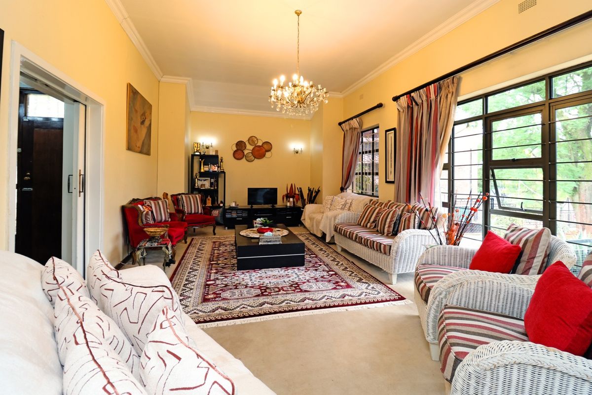A spacious living room featuring large windows with security bars, crown molding, and a central chandelier, offering a bright and comfortable living space.