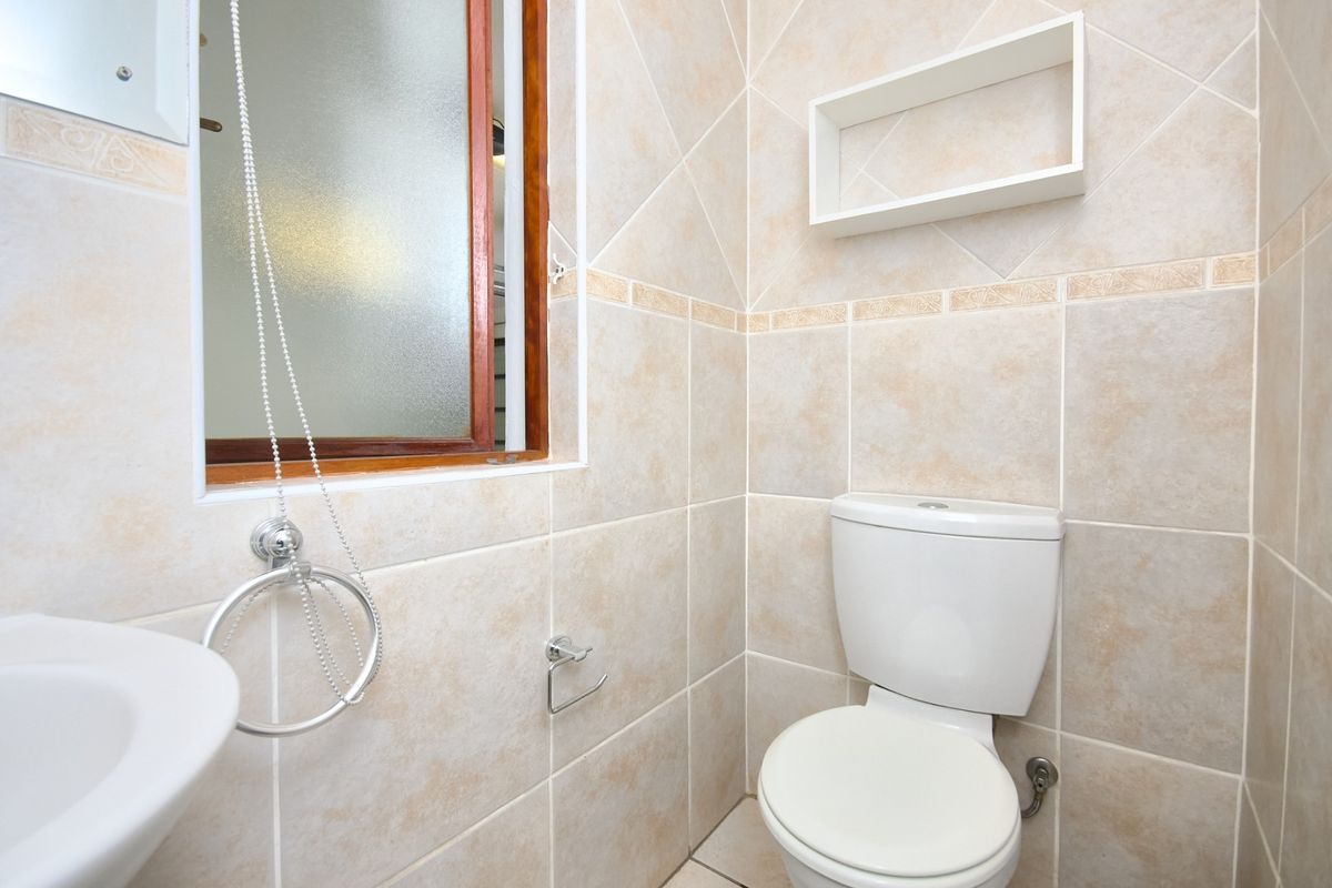A clean and functional bathroom featuring neutral wall tiles, a standard toilet, a washbasin, and a wall-mounted storage shelf.