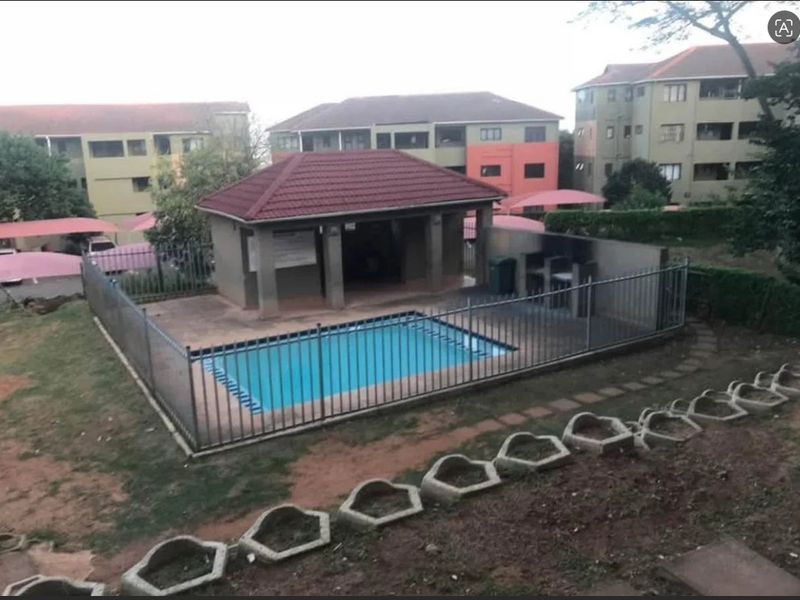 Communal swimming pool area featuring a covered gazebo, perimeter fencing, and paved surrounds within an apartment complex setting.
