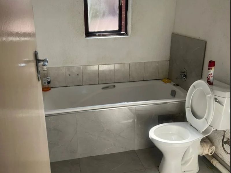 A functional bathroom featuring a built-in bathtub, tiled surround, and a standard toilet, with visible plumbing and neutral floor tiling.