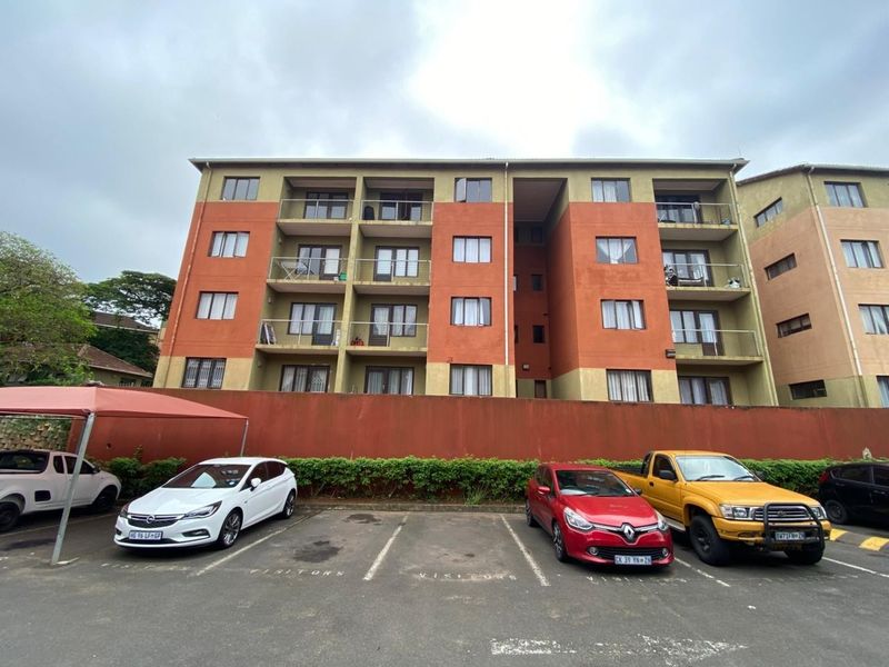 Exterior view of a multi-story apartment complex featuring private balconies and a paved visitor parking area with a covered carport structure.