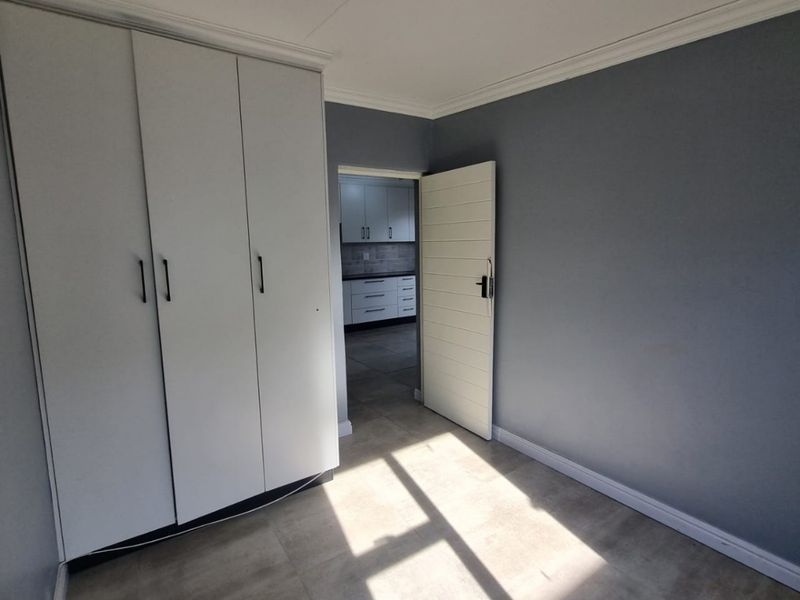 A bright, empty bedroom featuring neutral grey walls, tiled flooring, and built-in cupboards, with a view through to the kitchen area.