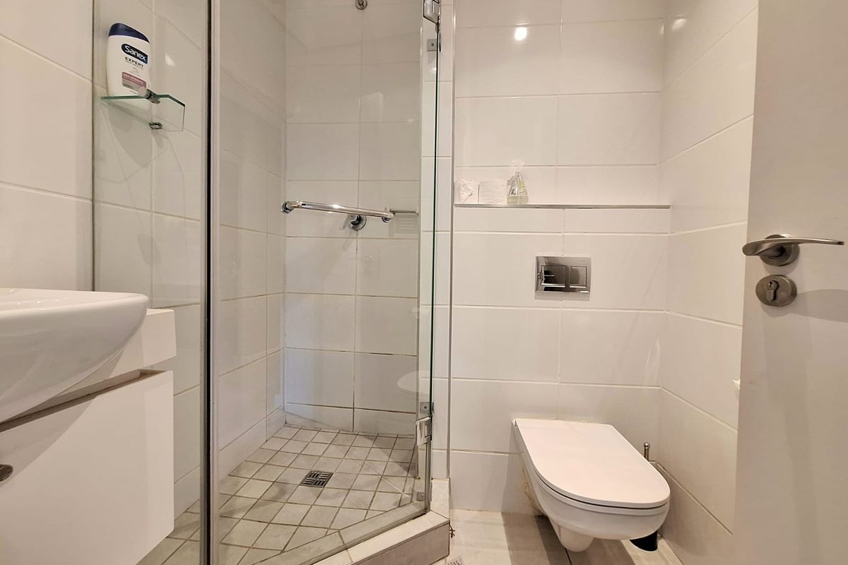 Compact modern bathroom featuring a glass-enclosed corner shower, wall-mounted toilet, and a sleek vanity basin with white tiled finishes.