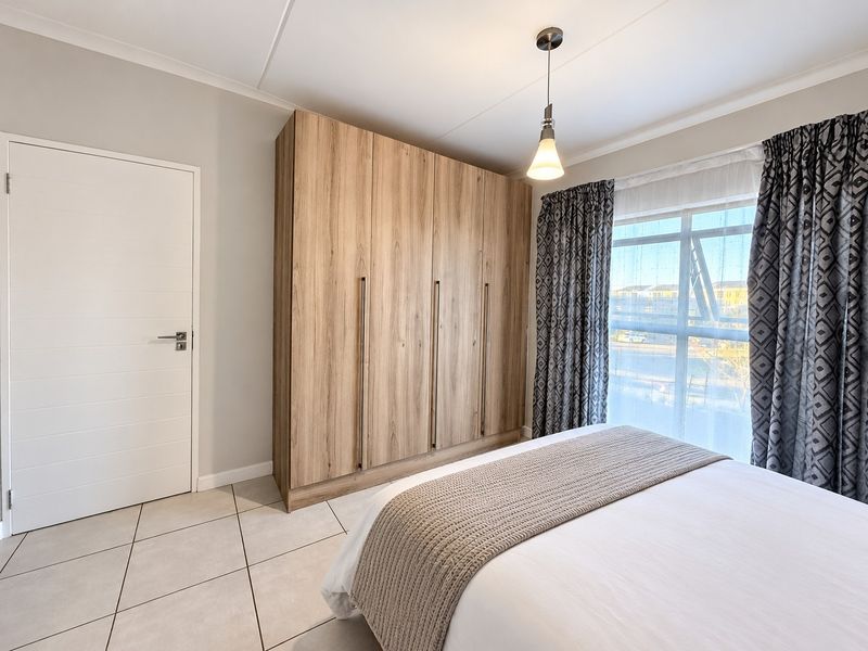A well-maintained bedroom featuring neutral tiled flooring, a modern built-in wooden wardrobe, and a large window with natural light.