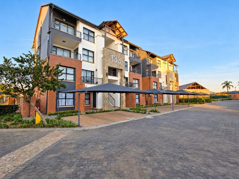 A modern multi-story apartment complex featuring brick and stone facade, private balconies, and covered parking bays within a well-maintained residential estate.