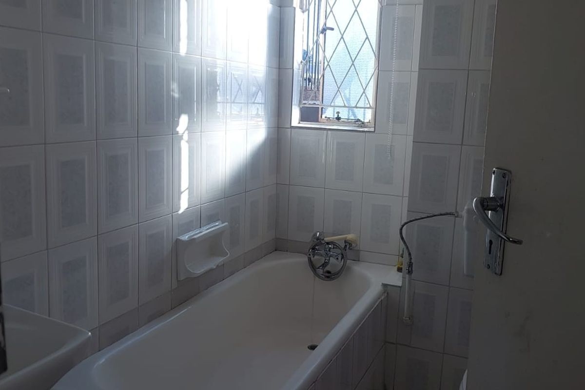 A functional bathroom featuring a standard bathtub, full-height wall tiling, and a window with security bars.