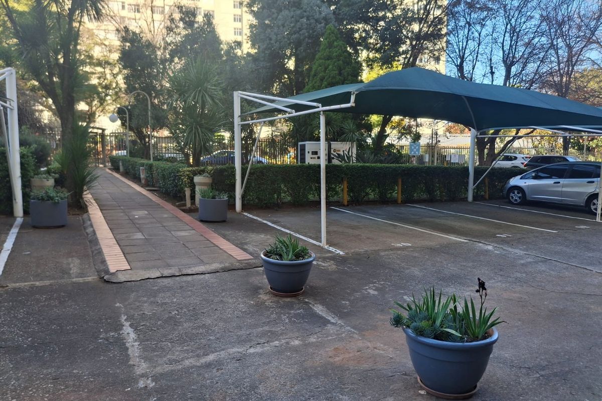 Outdoor parking area featuring a covered carport structure, paved walkway, and established perimeter hedging within a residential apartment complex.