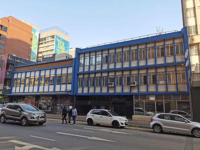 Street-level view of a multi-story commercial building facade featuring metal-framed windows and security gates, showing signs of age and weathering.