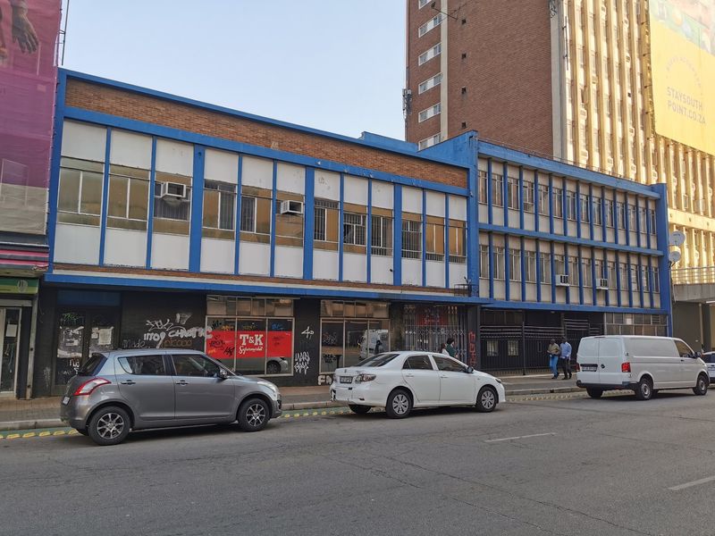 Street-level view of a multi-story commercial building featuring large display windows, security gates, and window-mounted air conditioning units.