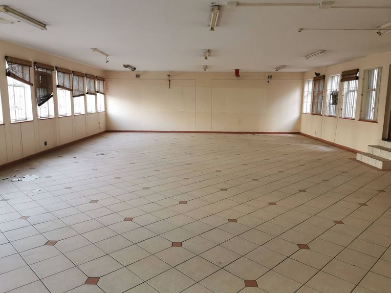 A large, vacant open-plan commercial space featuring tiled flooring, multiple windows with security bars, and overhead fluorescent lighting, suitable for various business configurations.