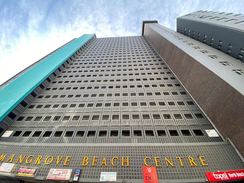 Low-angle exterior view of the Mangrove Beach Centre apartment building, showcasing its high-rise facade and ground-level commercial signage.