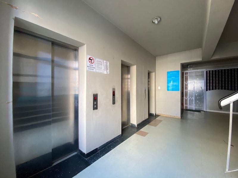 A communal apartment building hallway featuring multiple stainless steel elevator doors, tiled flooring, and access to a secure entrance area.