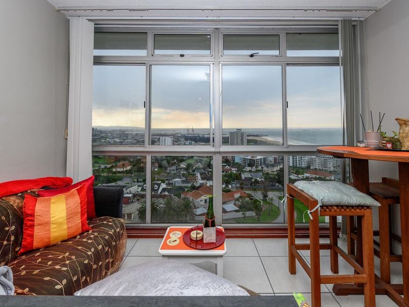 A living area featuring large aluminium-framed windows that provide expansive coastal and city views, complemented by neutral tiled flooring.