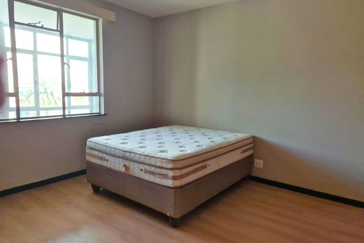 A simple bedroom featuring laminate flooring, neutral wall tones, and a large window providing natural light.