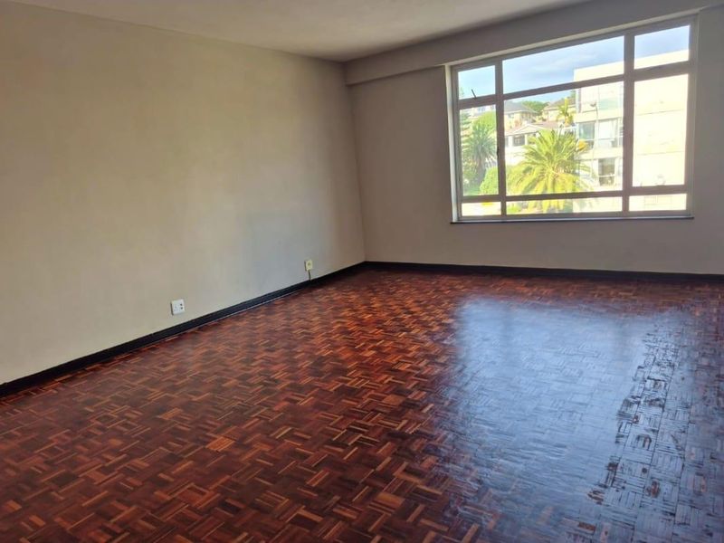 A spacious, empty living room featuring classic parquet flooring and a large window that provides ample natural light.
