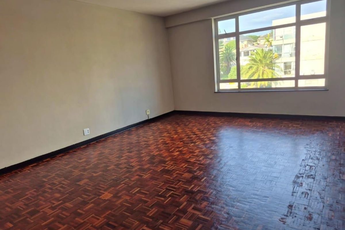 A spacious, empty living room featuring classic parquet flooring and a large window that provides ample natural light.