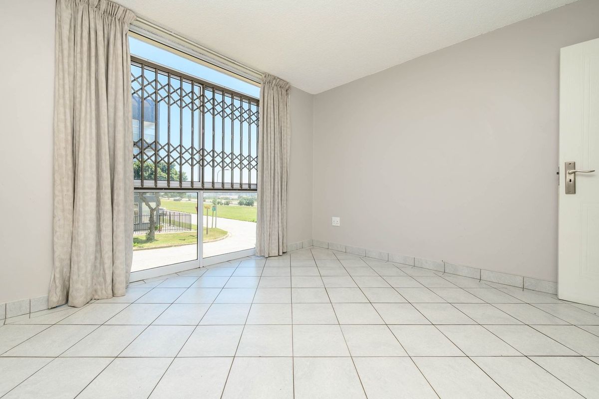 A bright, empty bedroom featuring neutral tiled flooring, a large window with a security gate, and clean wall finishes.
