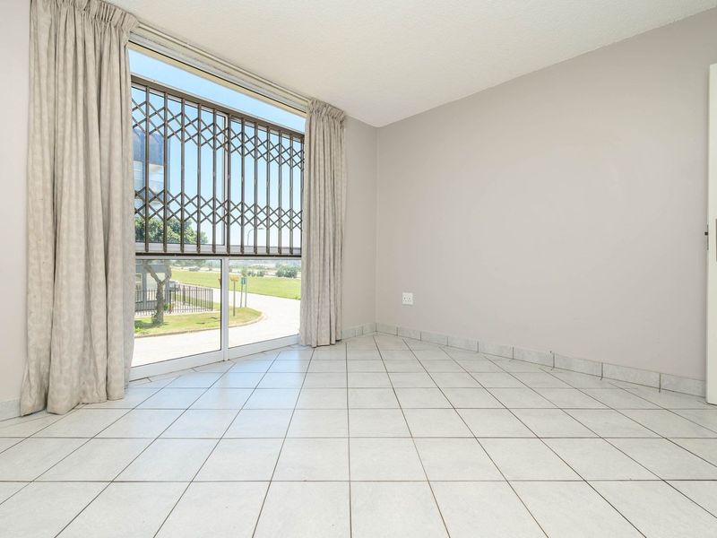 A bright, empty bedroom featuring neutral tiled flooring, a large window with a security gate, and clean wall finishes.