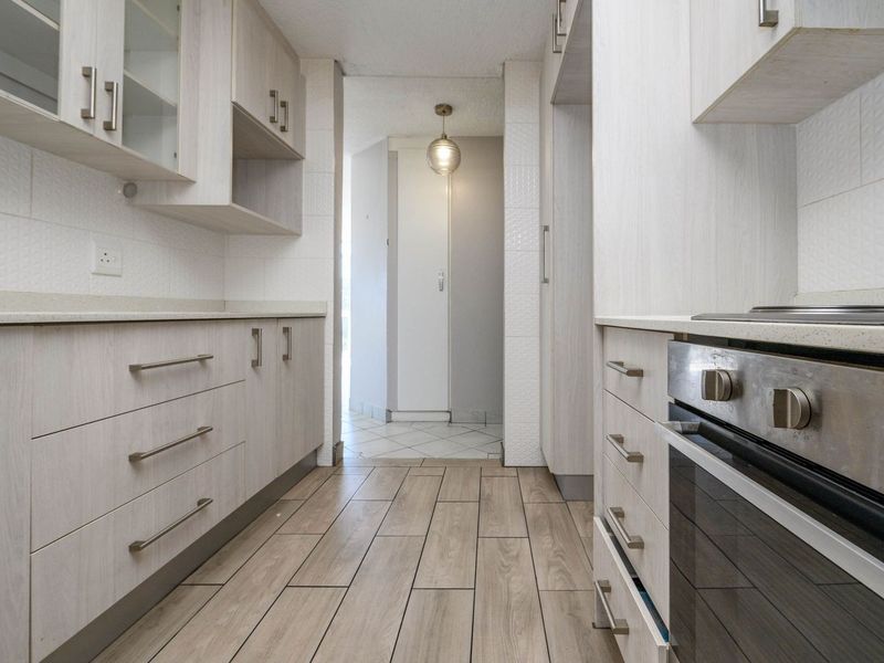 A clean, galley-style kitchen featuring light wood-finish cabinetry, stone countertops, built-in oven and hob, and modern wood-look floor tiles.