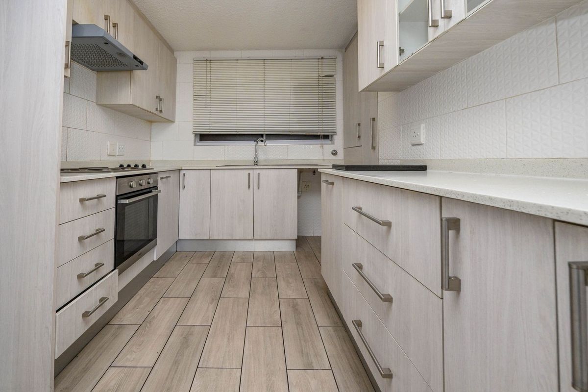 A clean, functional galley-style kitchen featuring light wood-finish cabinetry, stone countertops, integrated appliances, and modern wood-look floor tiles.