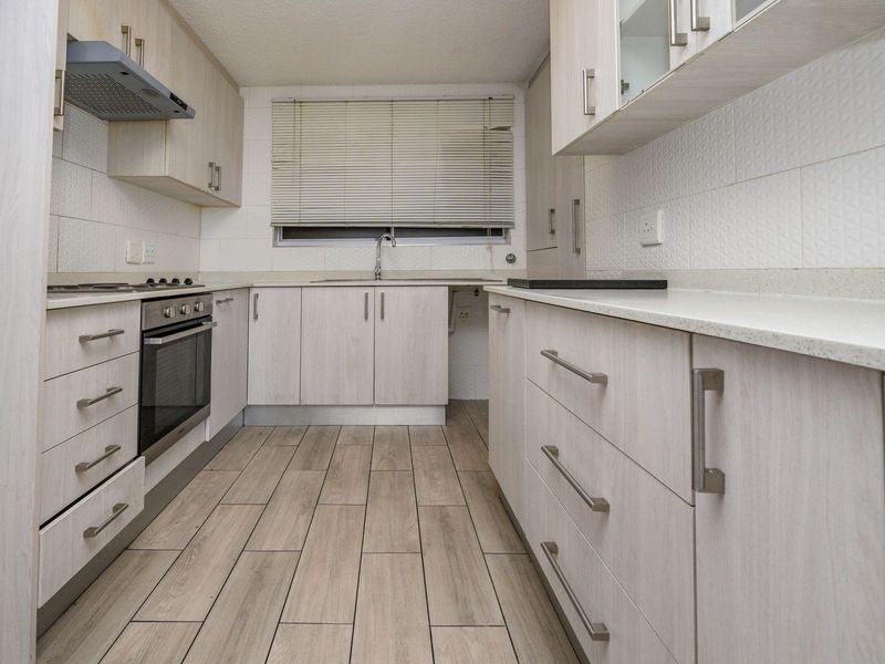 A clean, functional galley-style kitchen featuring light wood-finish cabinetry, stone countertops, integrated appliances, and modern wood-look floor tiles.