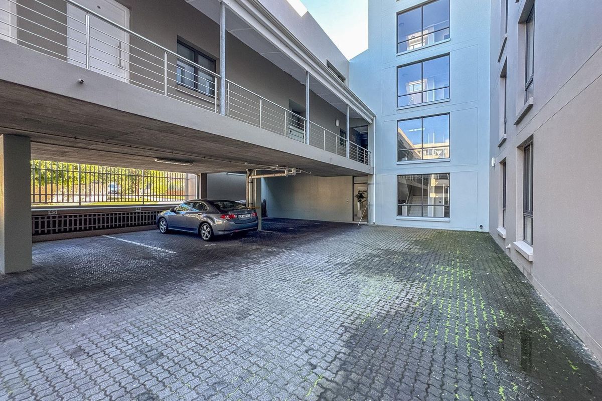 A view of the apartment complex exterior featuring a paved parking area, covered walkway, and multi-story building facade with large windows.
