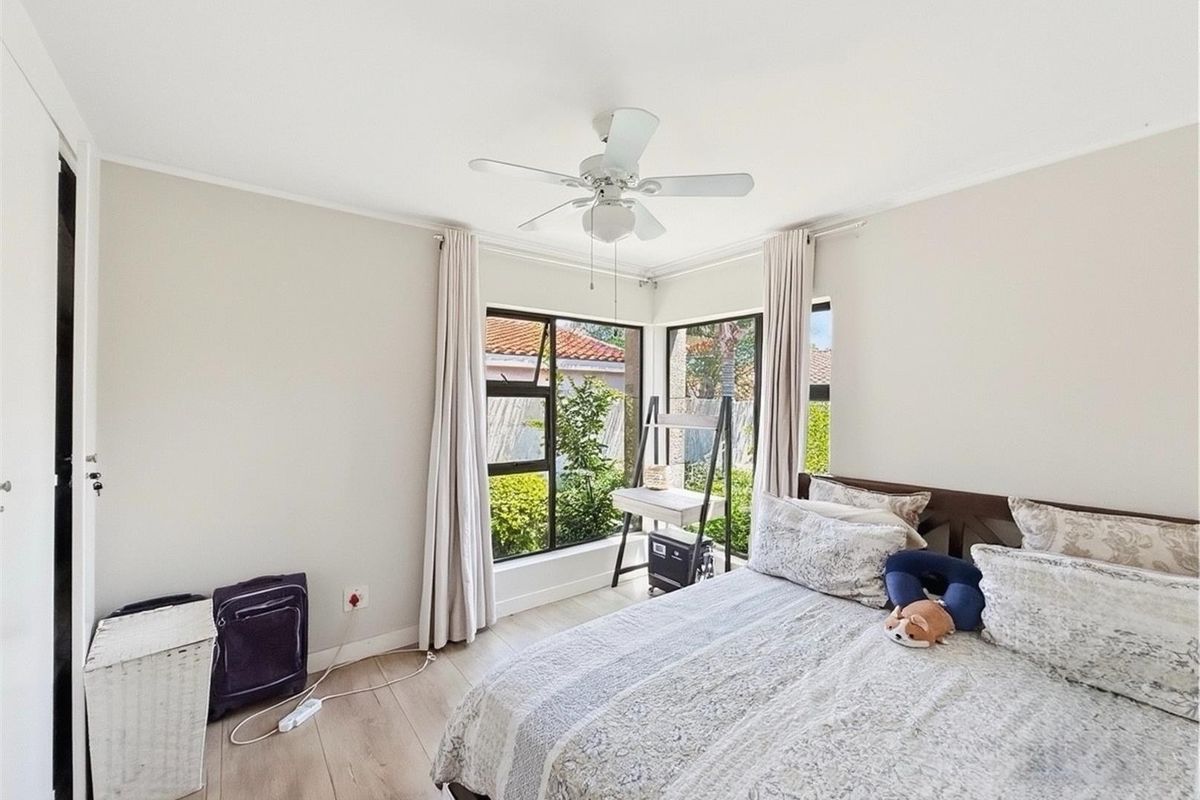 A bright bedroom featuring large corner windows, light wood-look flooring, and a ceiling fan, offering a pleasant view of the garden.