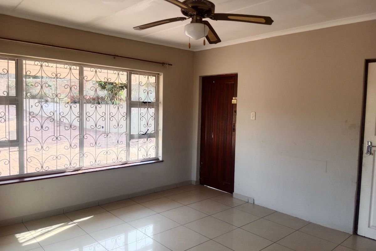 A bright, empty living room featuring tiled flooring, a ceiling fan, and large windows fitted with security bars.