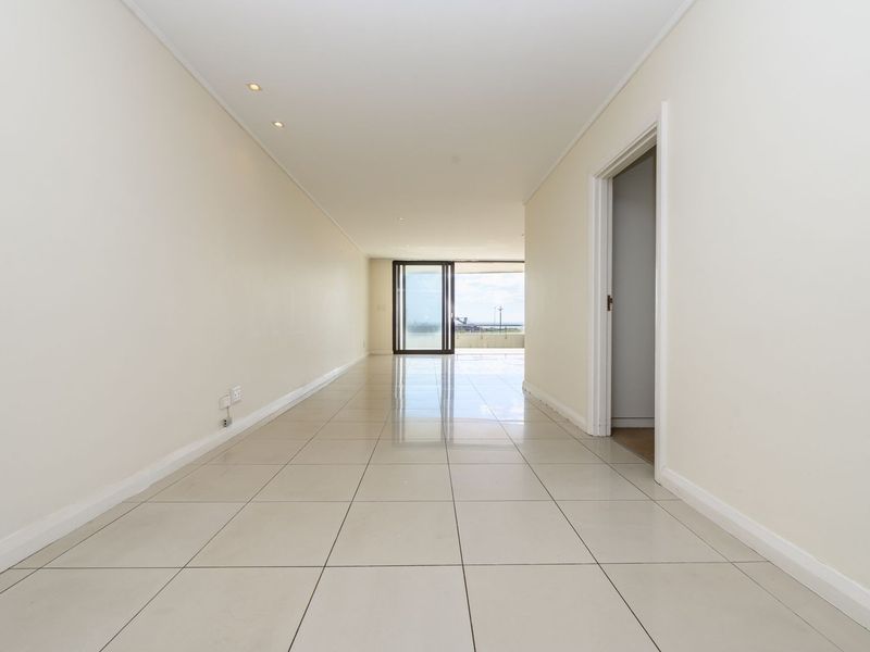 A bright, empty hallway featuring neutral tiled flooring, recessed lighting, and a sliding glass door leading to an outdoor area.