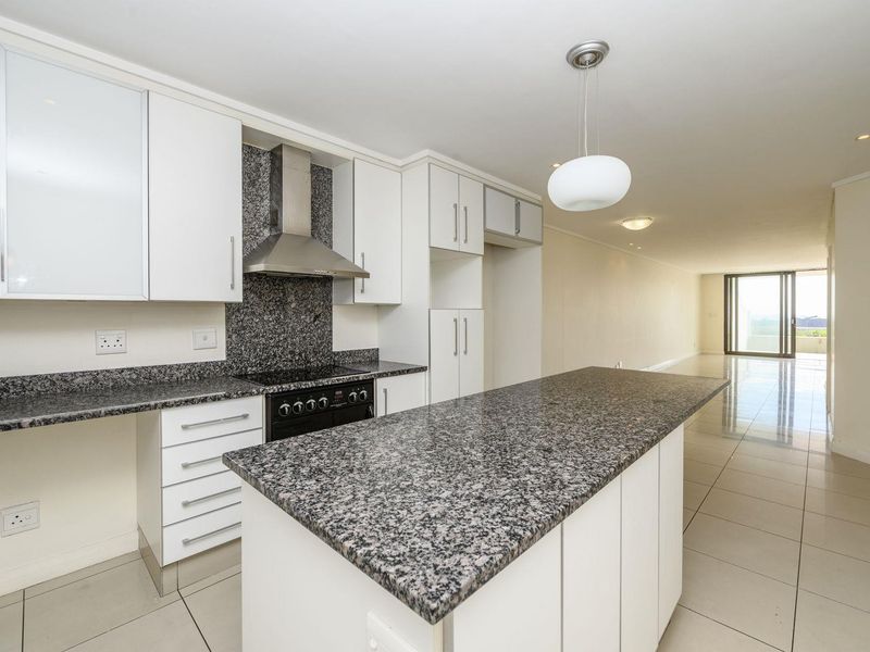 Modern open-plan kitchen featuring granite countertops, ample white cabinetry, and a central island, flowing seamlessly into a spacious living area with tiled floors.
