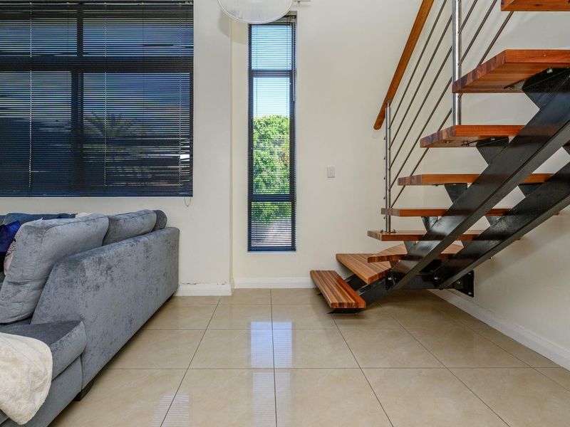 Modern living area featuring a striking floating staircase with timber treads, stainless steel balustrades, large format floor tiles, and tall windows with venetian blinds.