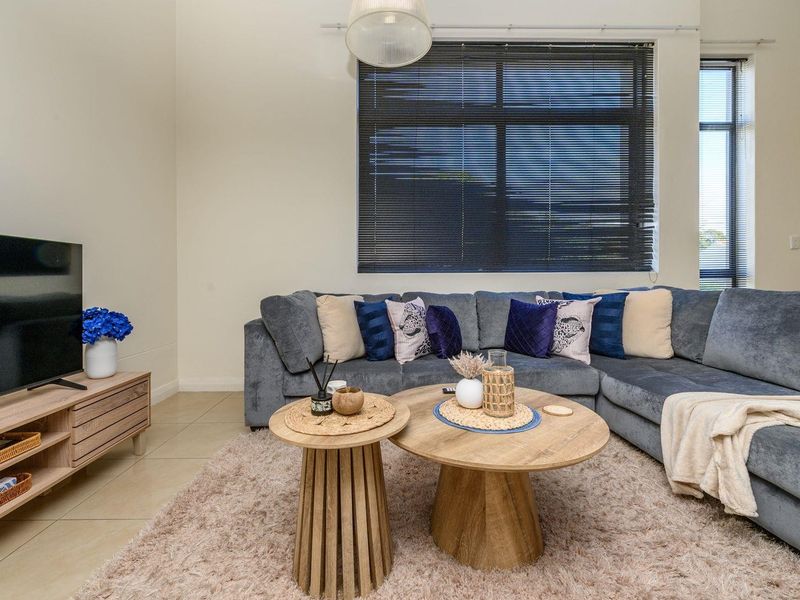 A well-maintained living room featuring neutral tiled flooring, large windows with venetian blinds, and a modern pendant light fixture.