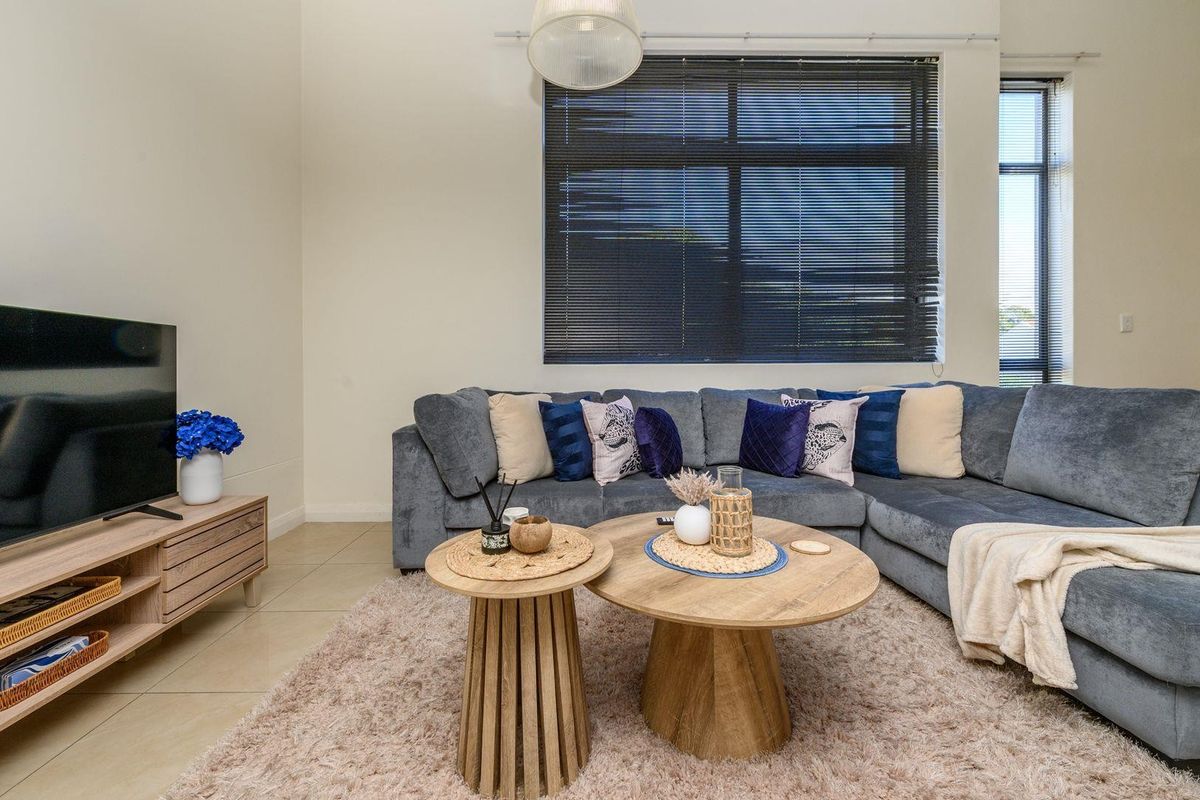 A well-maintained living room featuring neutral tiled flooring, large windows with venetian blinds, and a modern pendant light fixture.