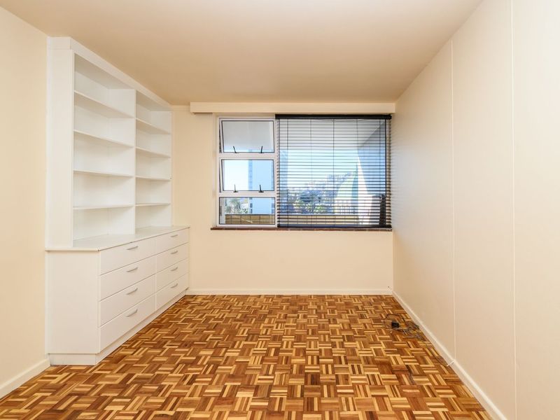 A bright, empty bedroom featuring classic parquet flooring, built-in white cabinetry with shelving and drawers, and a window with fitted blinds.