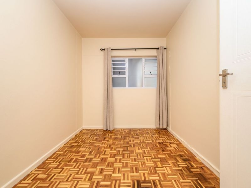 A bright, empty bedroom featuring well-maintained parquet flooring and a louvered window with a curtain rod.