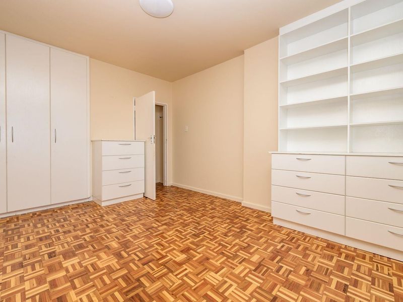 A bright bedroom featuring classic parquet flooring, extensive built-in cabinetry, and integrated shelving units, offering ample storage space in a clean, neutral setting.