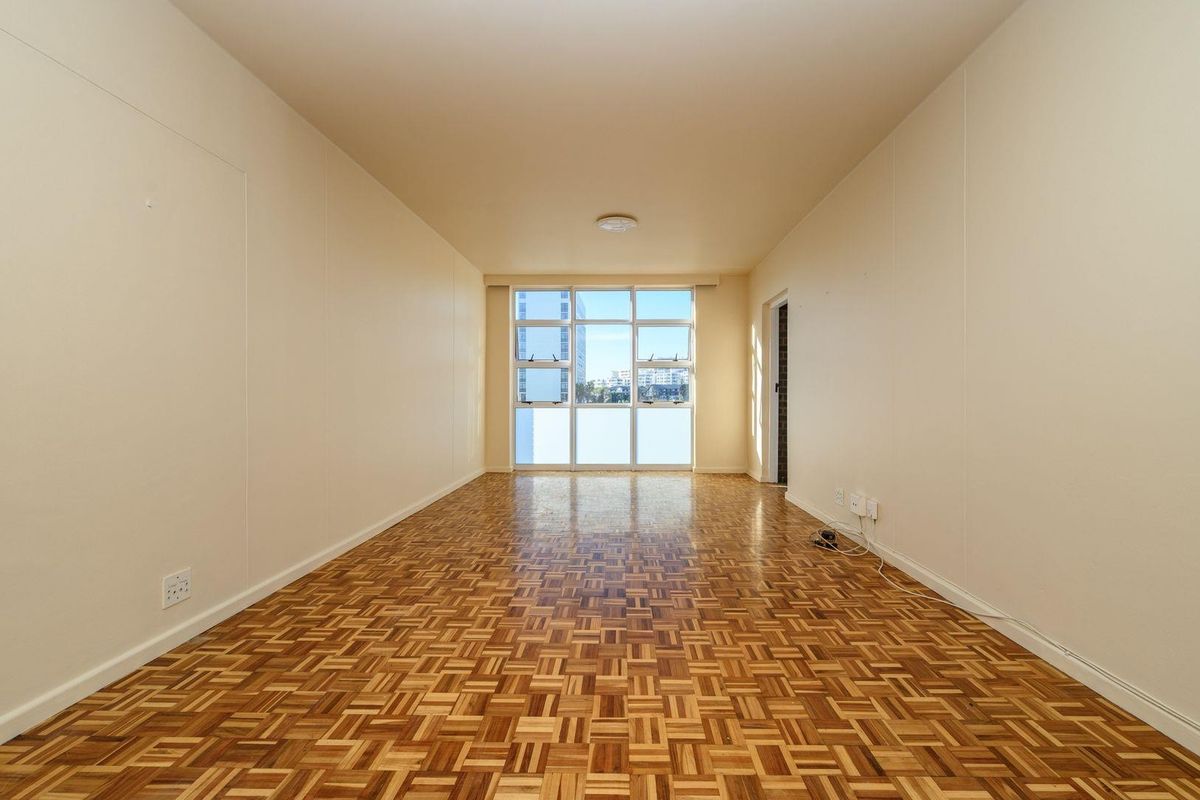 A bright, empty living room featuring classic parquet flooring, neutral walls, and a large window providing natural light.