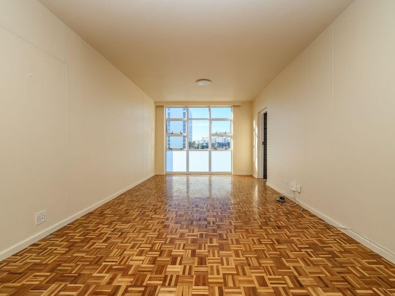 A bright, empty living room featuring classic parquet flooring, neutral walls, and a large window providing natural light.