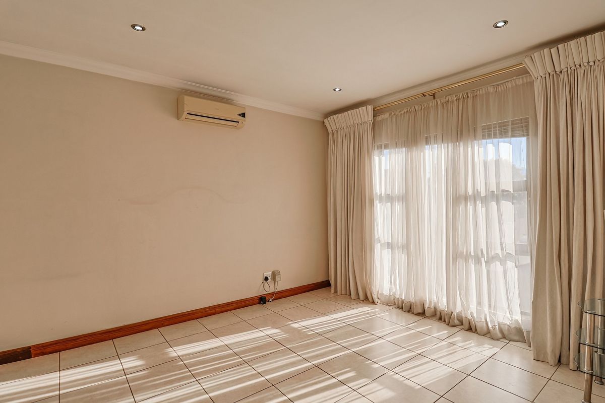 A bright, empty bedroom featuring tiled flooring, recessed ceiling lighting, and a wall-mounted air conditioning unit with large windows covered by curtains.