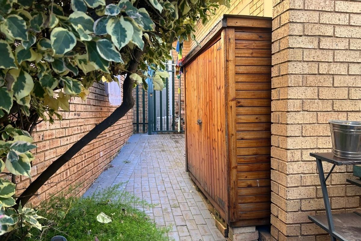 A side passage of the townhouse featuring brick paving, a face brick wall, and a built-in wooden storage shed leading to a security gate.