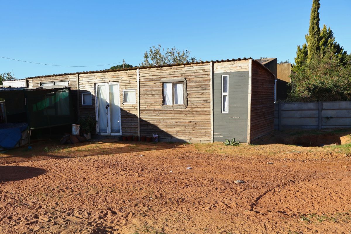 A detached timber-clad structure featuring a simple facade with double doors and windows, situated on an unlandscaped dirt plot.