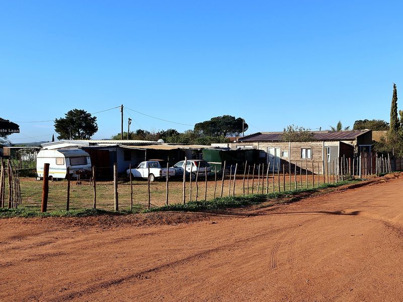 Front view of a property featuring timber-clad structures with corrugated metal roofing, situated along an unpaved road with basic perimeter fencing.