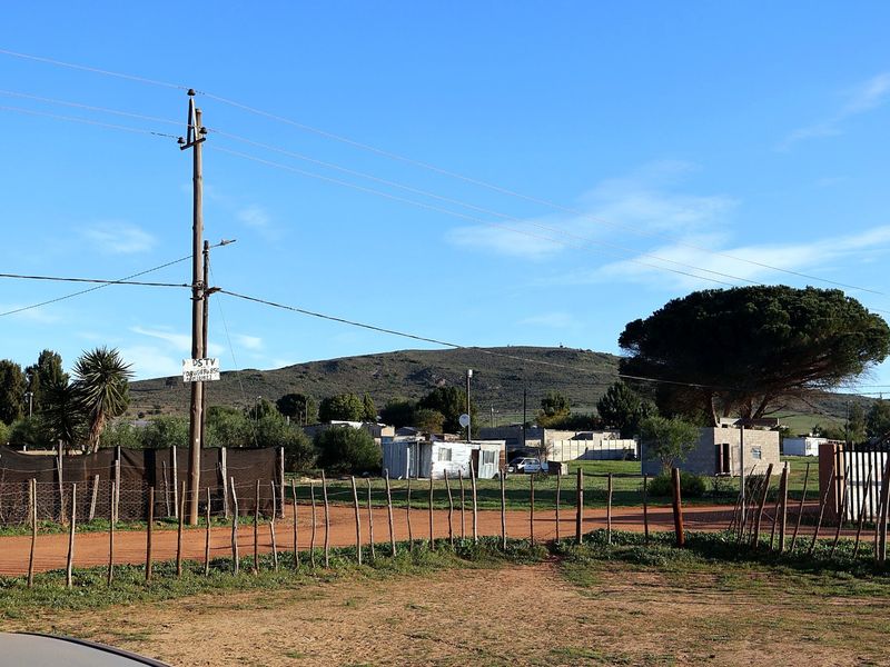 A view of the surrounding neighbourhood and street approach, featuring a dirt road, utility infrastructure, and open land with distant hills.