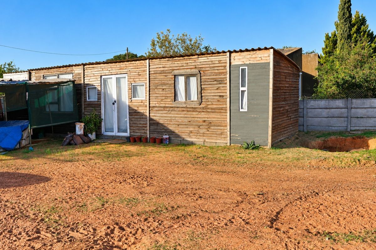 A detached timber-clad structure featuring double glass doors and casement windows, situated on an unpaved plot with a concrete boundary wall.