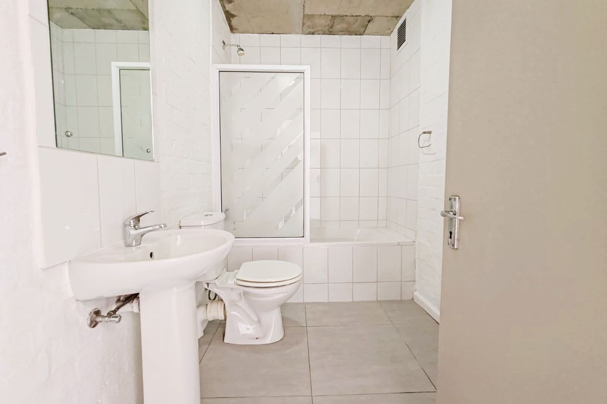 A functional bathroom featuring a pedestal basin, toilet, and a bathtub with a shower screen, finished with white wall tiles and grey floor tiles.