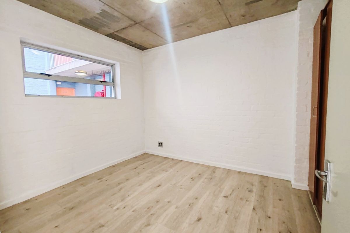 A bright, empty bedroom featuring modern laminate flooring, painted brick walls, and an exposed concrete ceiling with a built-in cupboard.