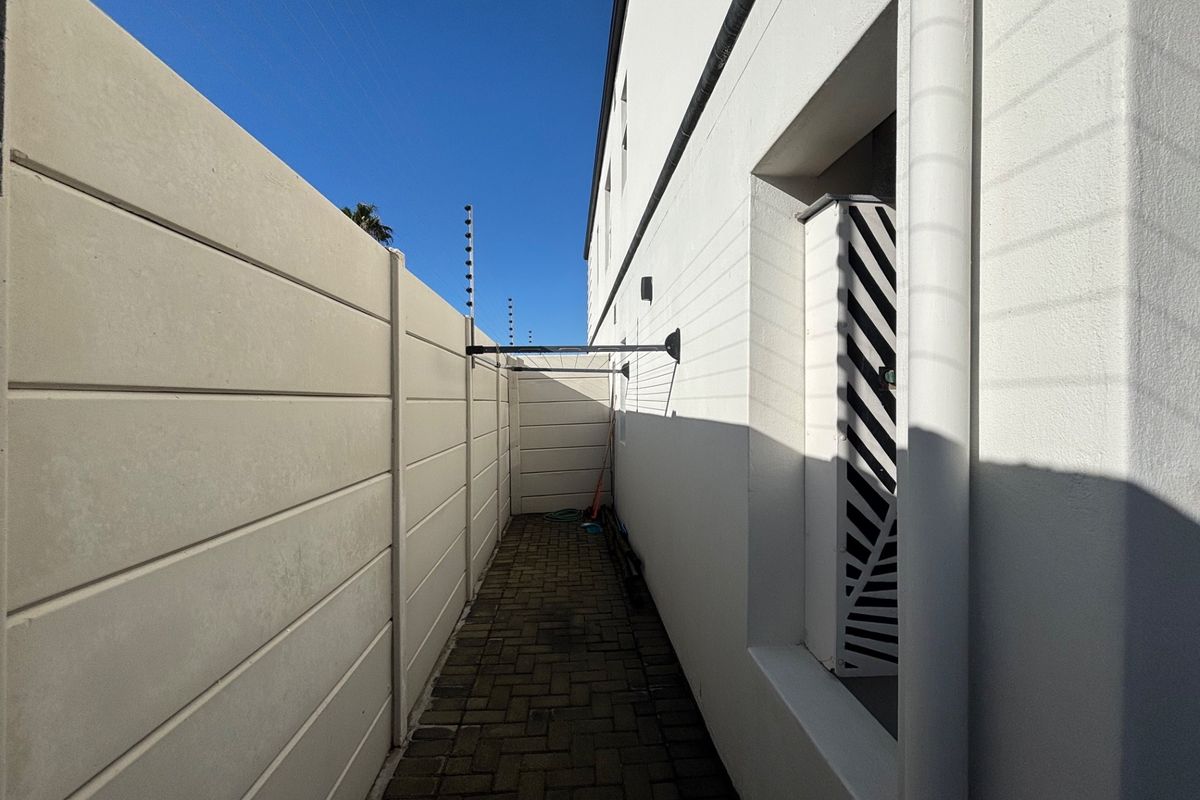 A narrow side passage featuring paved flooring, a high boundary wall with electric fencing, and a wall-mounted washing line.