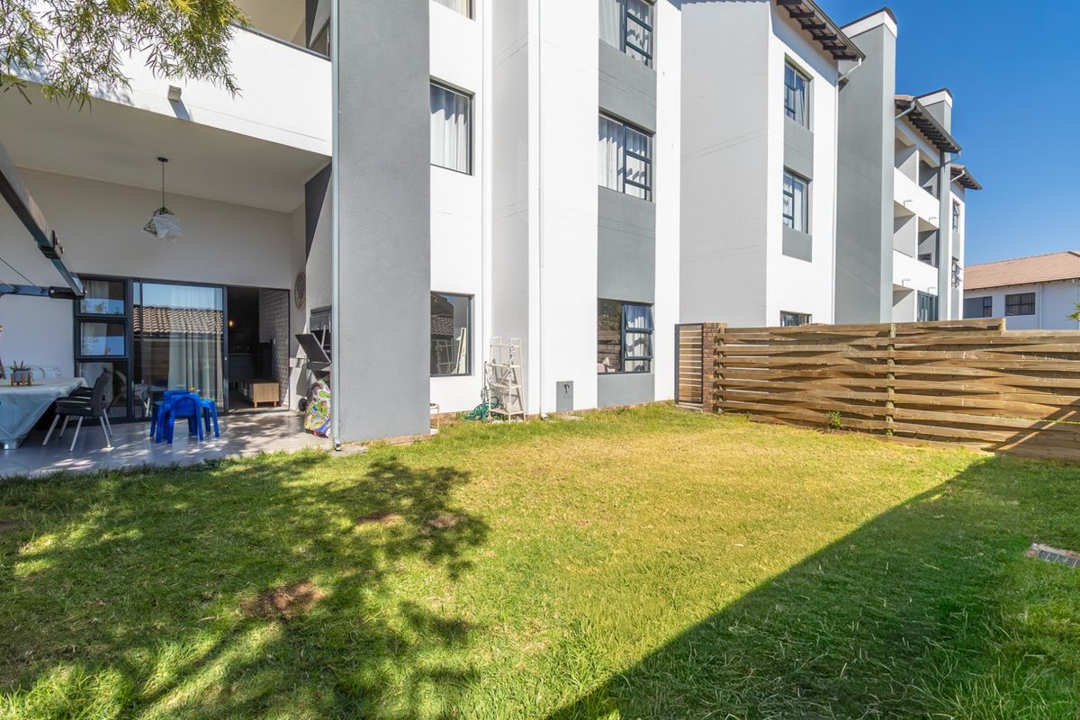 A ground-floor apartment featuring a private garden, a covered patio area, and modern multi-story building architecture with sliding glass doors.